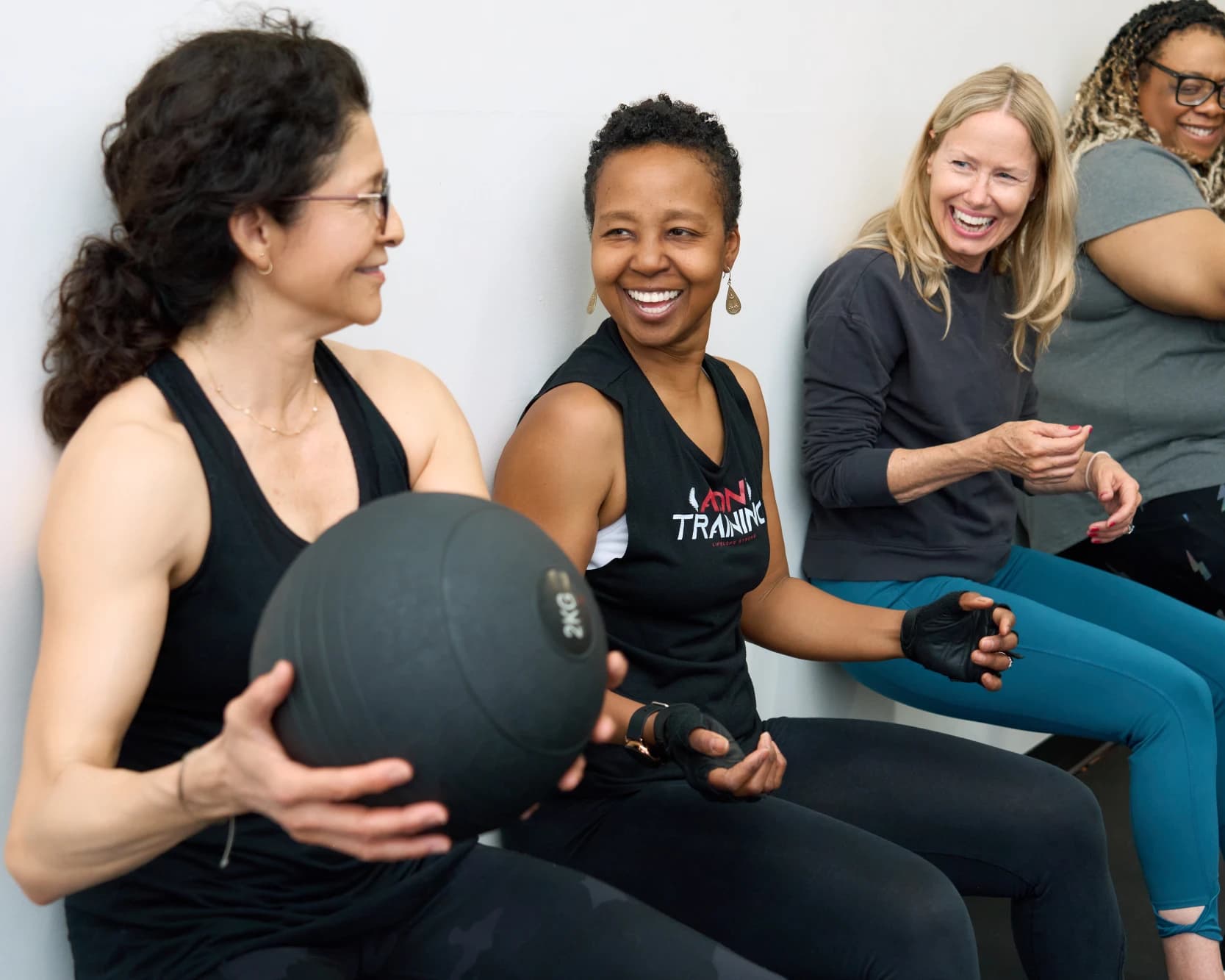 Three members sit against the studio wall laughing, one holding a medicine ball, between sets.