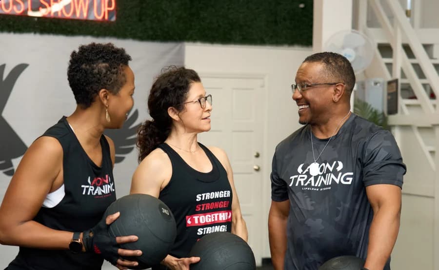 Two members holding medicine balls talk with a coach in front of the Just Show Up wall.
